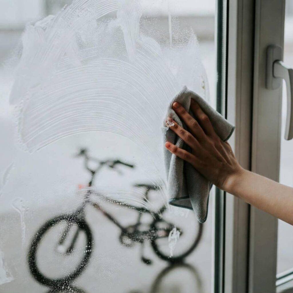Hand smears microfiber streaks on window; kid’s bike abandoned in background.