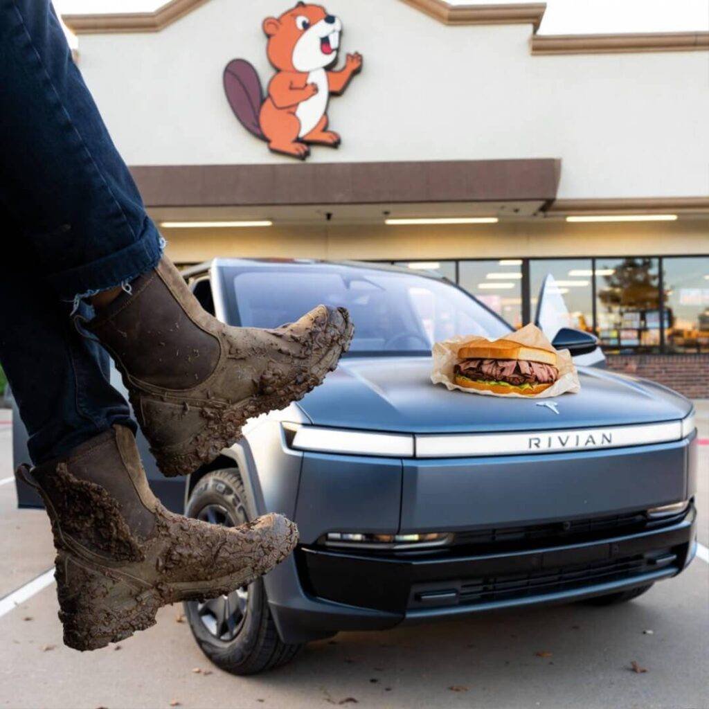 Muddy boots dangle from Rivian at Buc-ee’s, half brisket sandwich on hood.