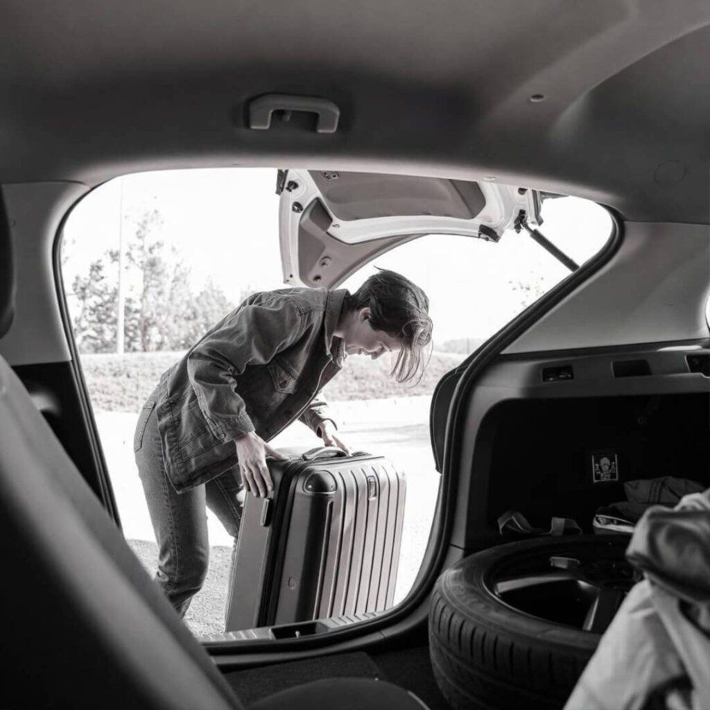 Person awkwardly loading suitcase into rental car, side mirror reflection.