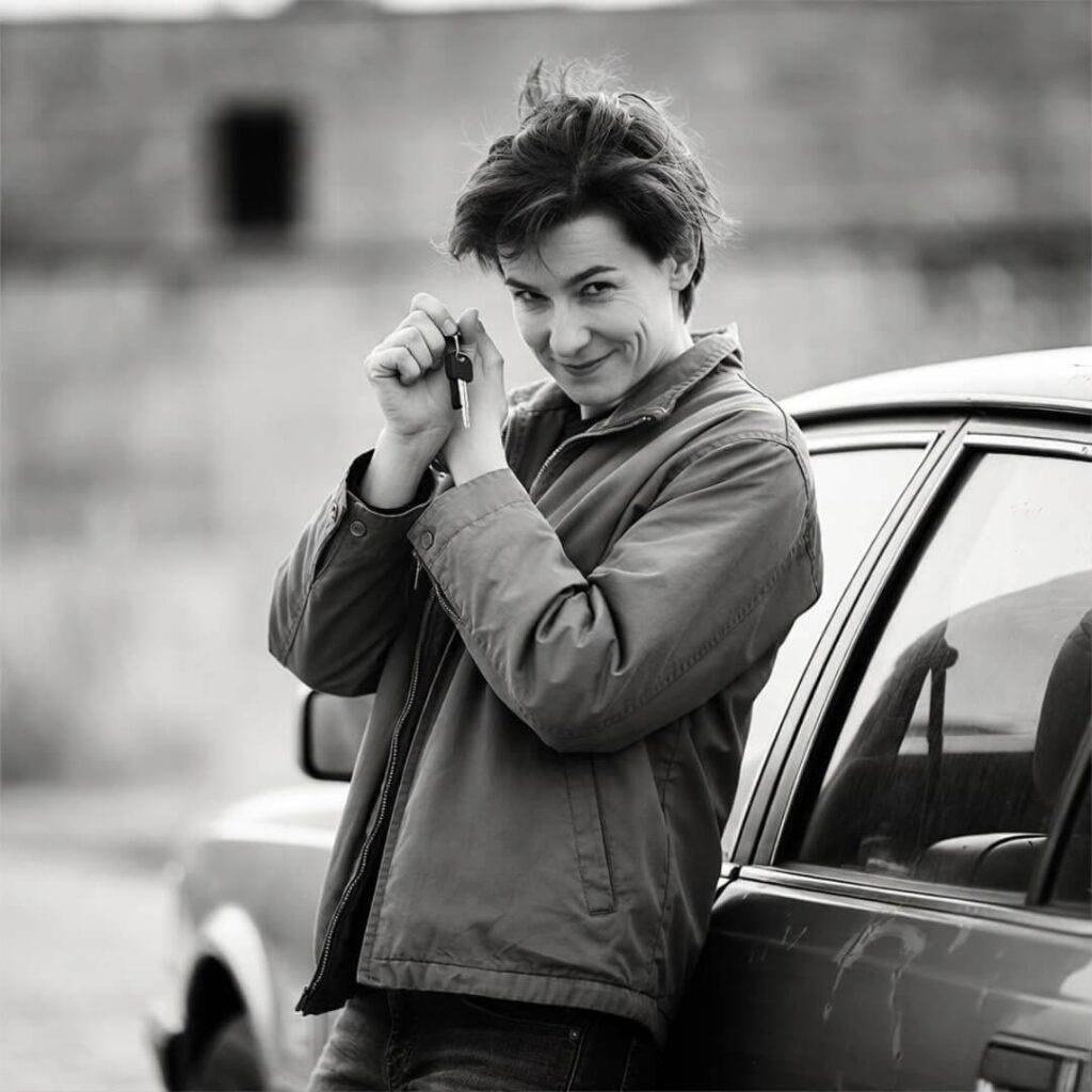 Person fumbling with rental car key, awkward grin, leaning on beat-up sedan.