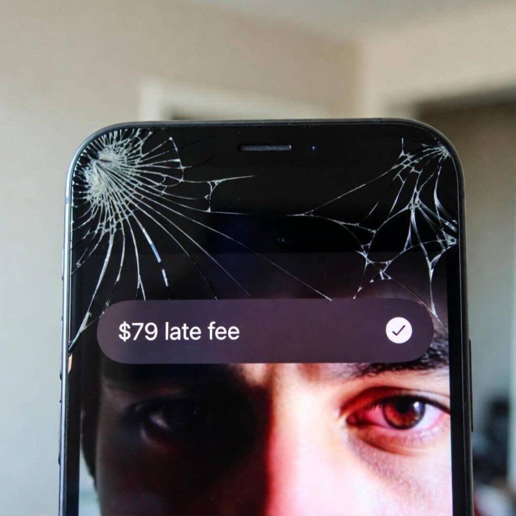 Cracked iPhone screen shows "$79 late fee" alert, bloodshot eye reflected.