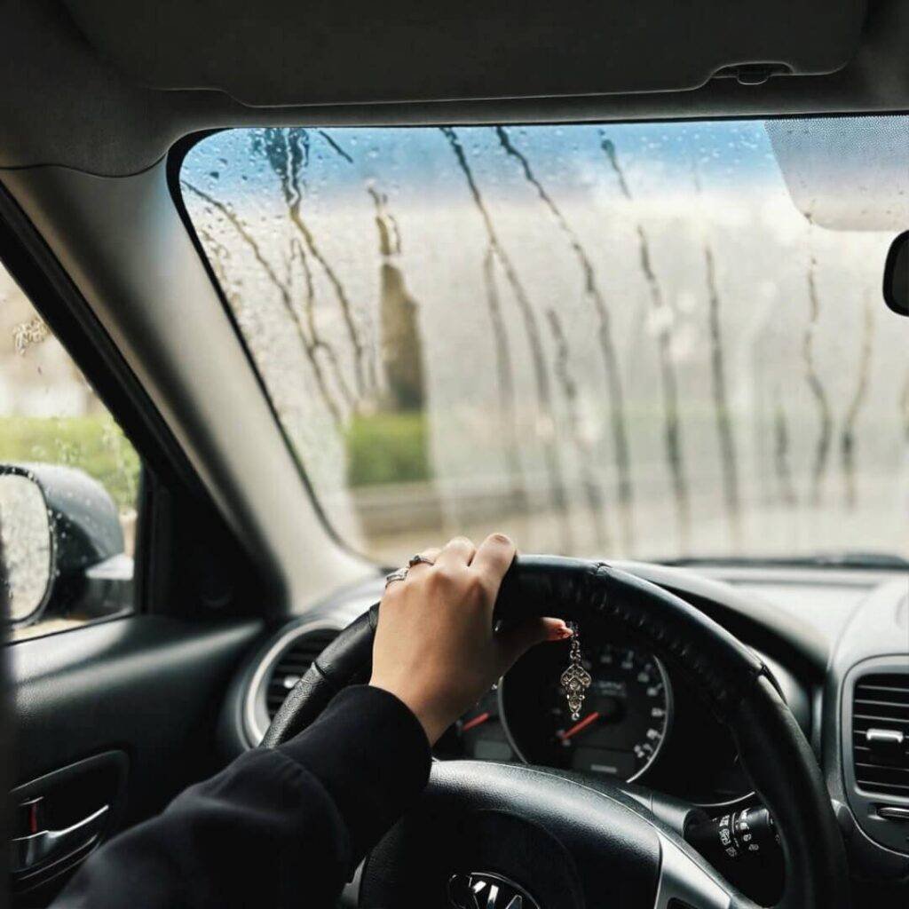 POV selfie: chipped nails grip Kia Soul wheel, rain streaks windshield.