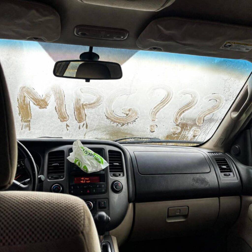 POV inside car: "MPG???" on foggy windshield, 48.2 mpg dash, Taco Bell wrapper on vent.