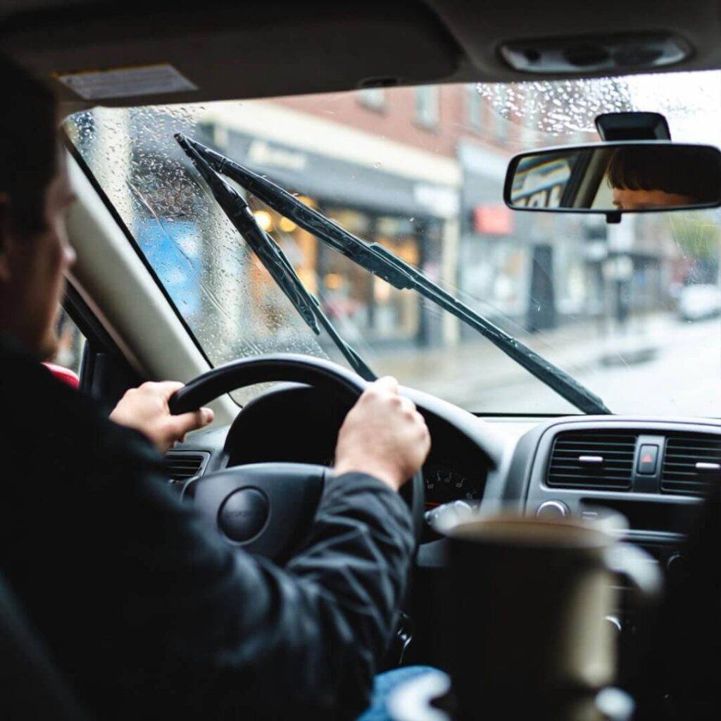 Driver's view: rainy Seattle street, wipers blur "SOLD" sign in mirror, coffee mug tipping.