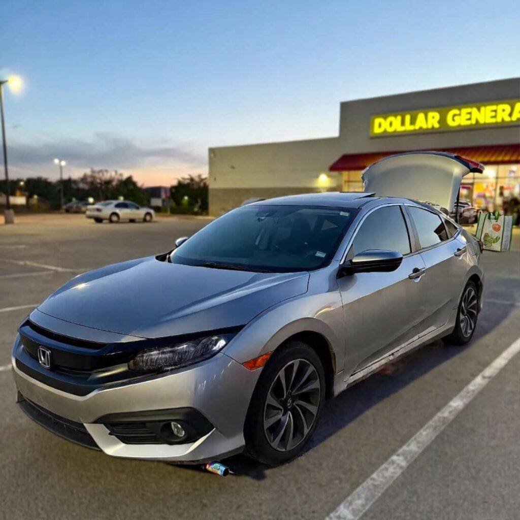 Low-angle silver Honda Civic at dusk, burnt headlight, groceries spilling, straw in tailpipe.