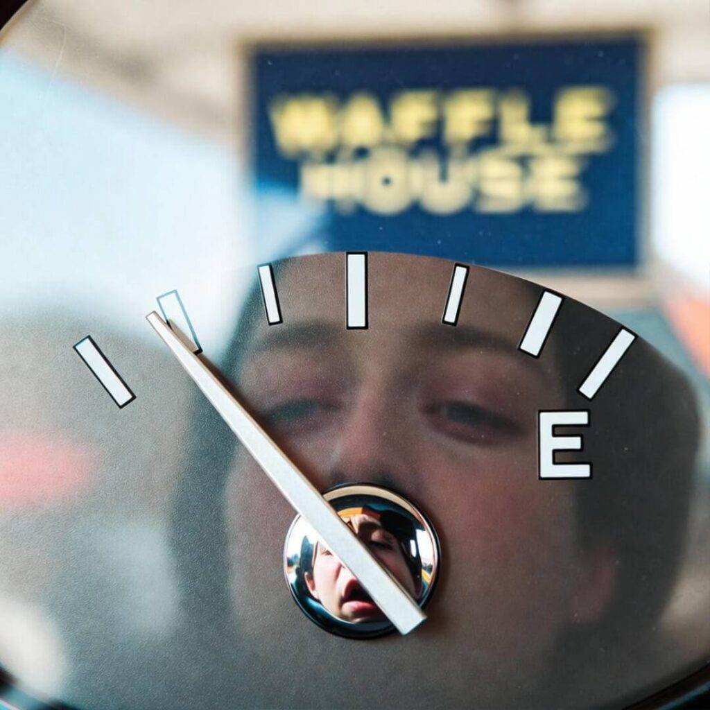 Fuel gauge near E, exhausted face reflected, blurred Waffle House sign behind.