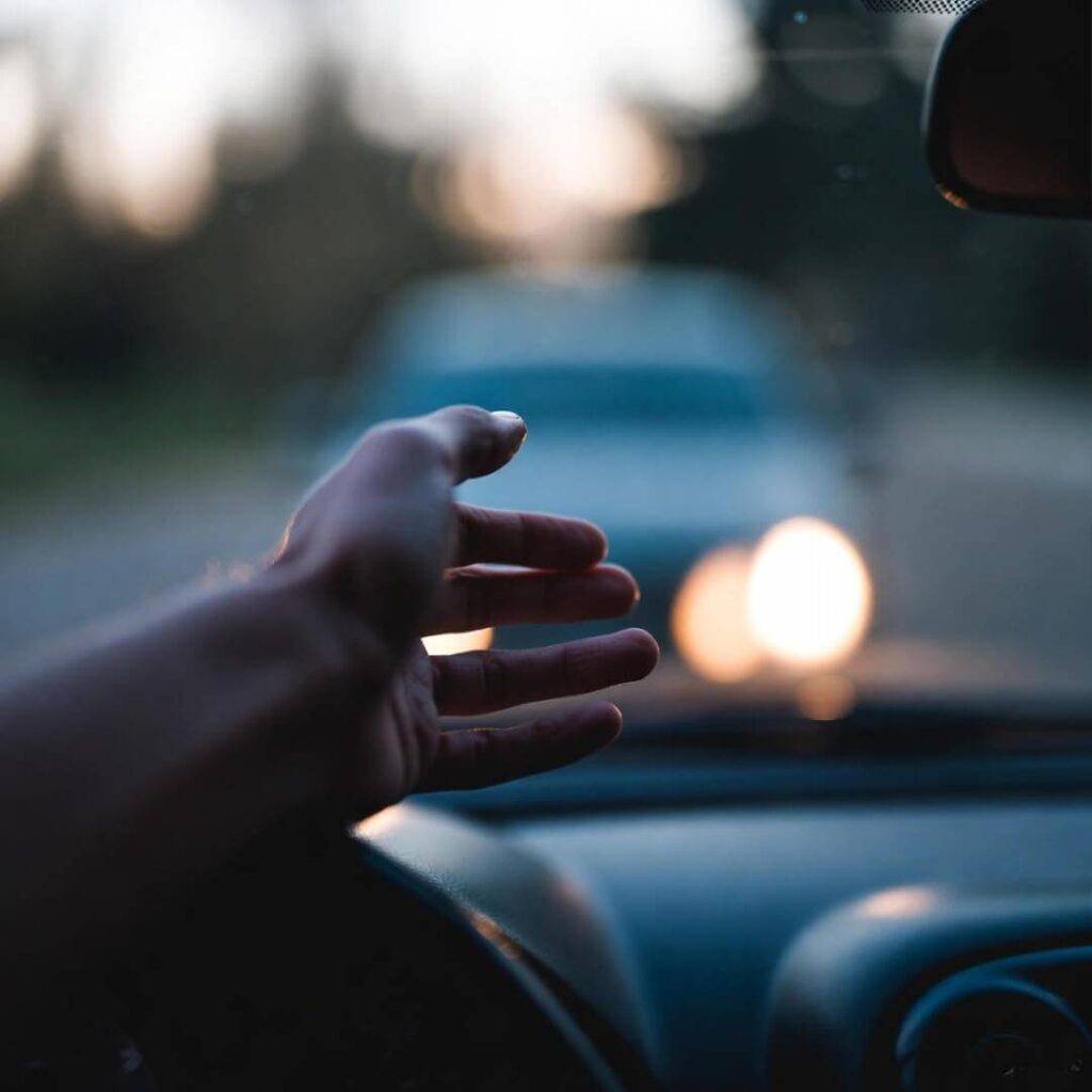 Hand hovering over dim headlights at twilight, low-angle from steering wheel.