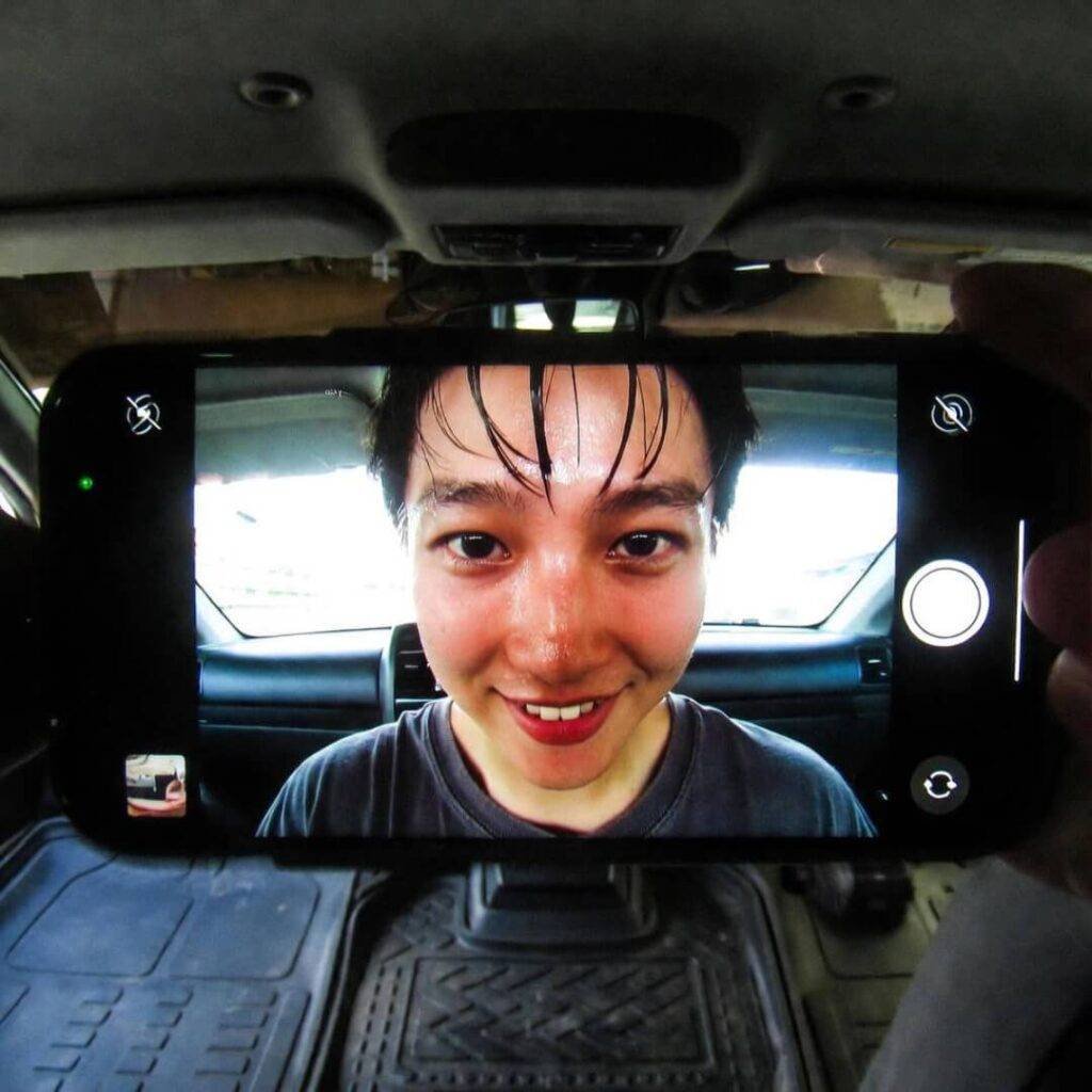 Sweaty grin in grainy phone selfie, lit by glowing screen from passenger footwell.