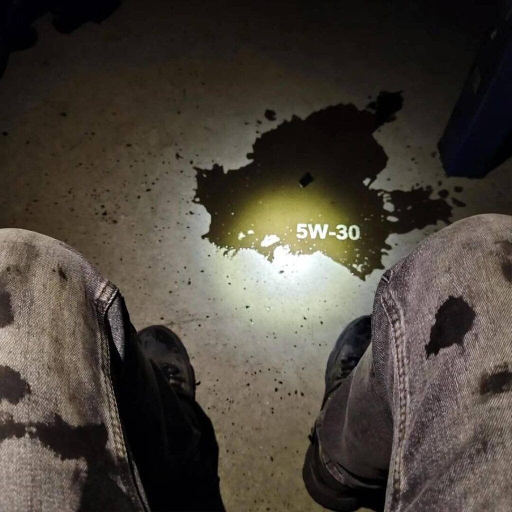 POV: Oil-stained jeans above 5W-30 puddle, lit by phone flashlight.