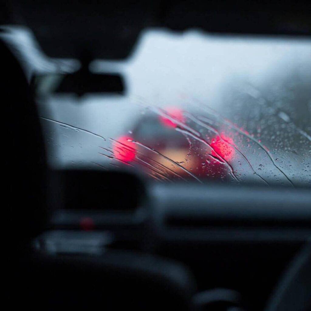 Blurry backseat view: rain streaks window, red taillights bleed into fog.