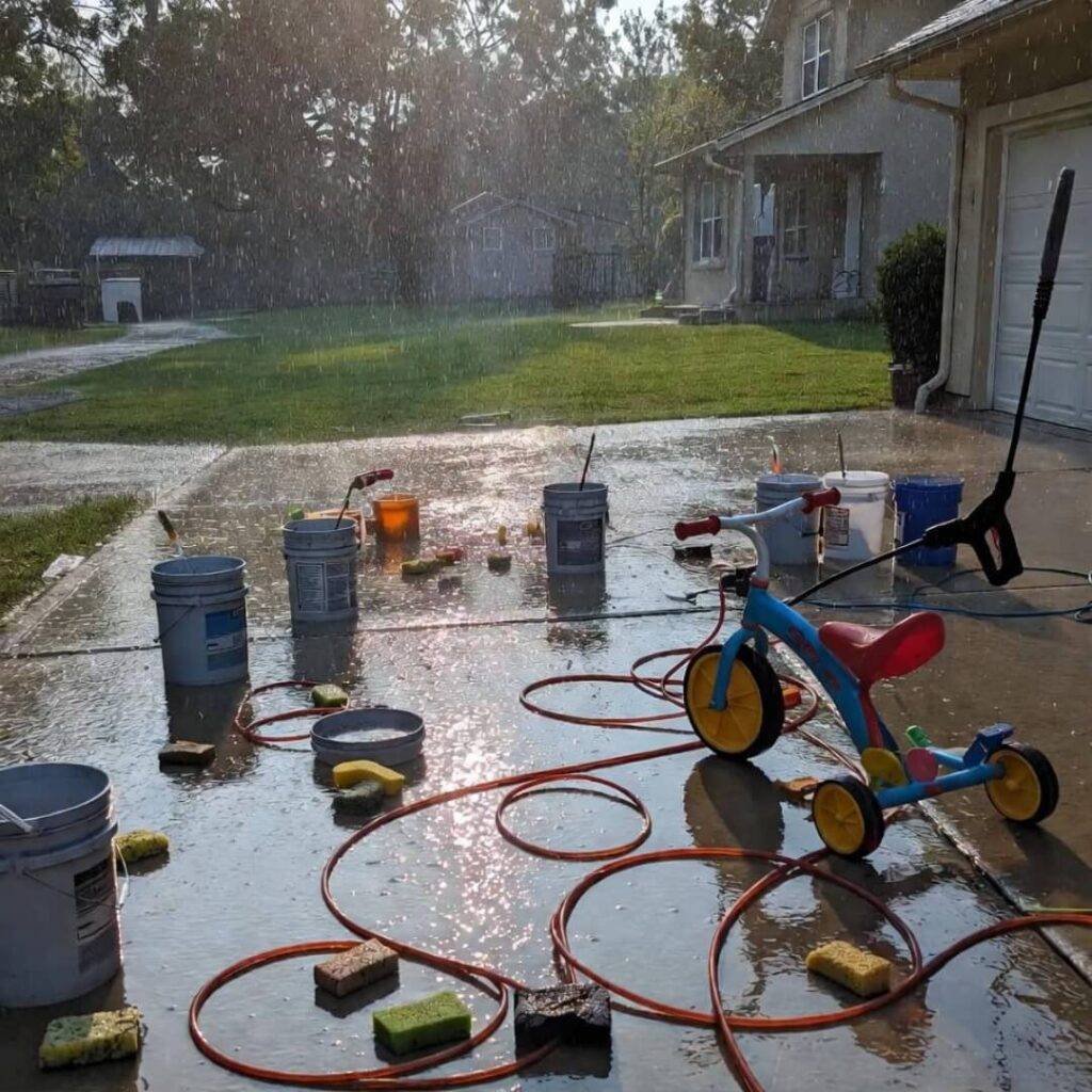 Rain-soaked driveway: buckets, cords, tricycle tangled in hose, sparkling mess.