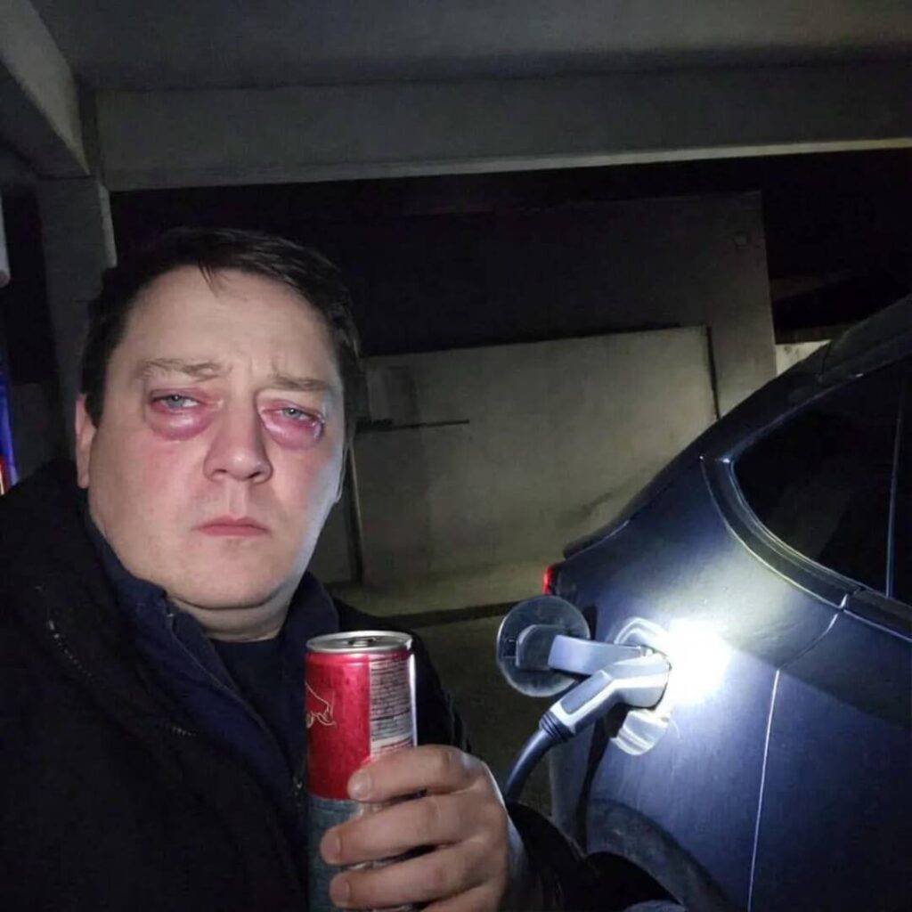 Grainy 2am selfie: bloodshot eyes, Red Bull, flashlight on Tesla charging port.