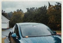 Electric Cars: Myths vs. Reality in 2025 Rain-soaked teal Tesla Model Y with coffee cup, autumn leaf, and skeptical reflection in puddle.
